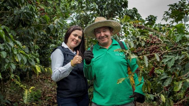A woman in a black vest and a man in a green shirt and straw hat pose in a field, surrounded by coffee plants.   From 2015 through 2022, Chemonics International implemented the Rural Finance Initiative (RFI) project. Funded by USAID, RFI worked with local financial institutions in Colombia to expand financial inclusion for Venezuelan migrants in urban areas and for Colombians in rural areas. Photo courtesy of RFI.
